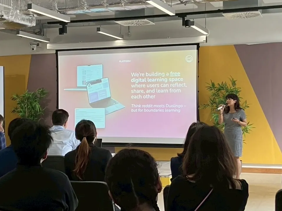 Cream speaking to a seated audience, beside a slide reading “We’re building a free digital learning space”